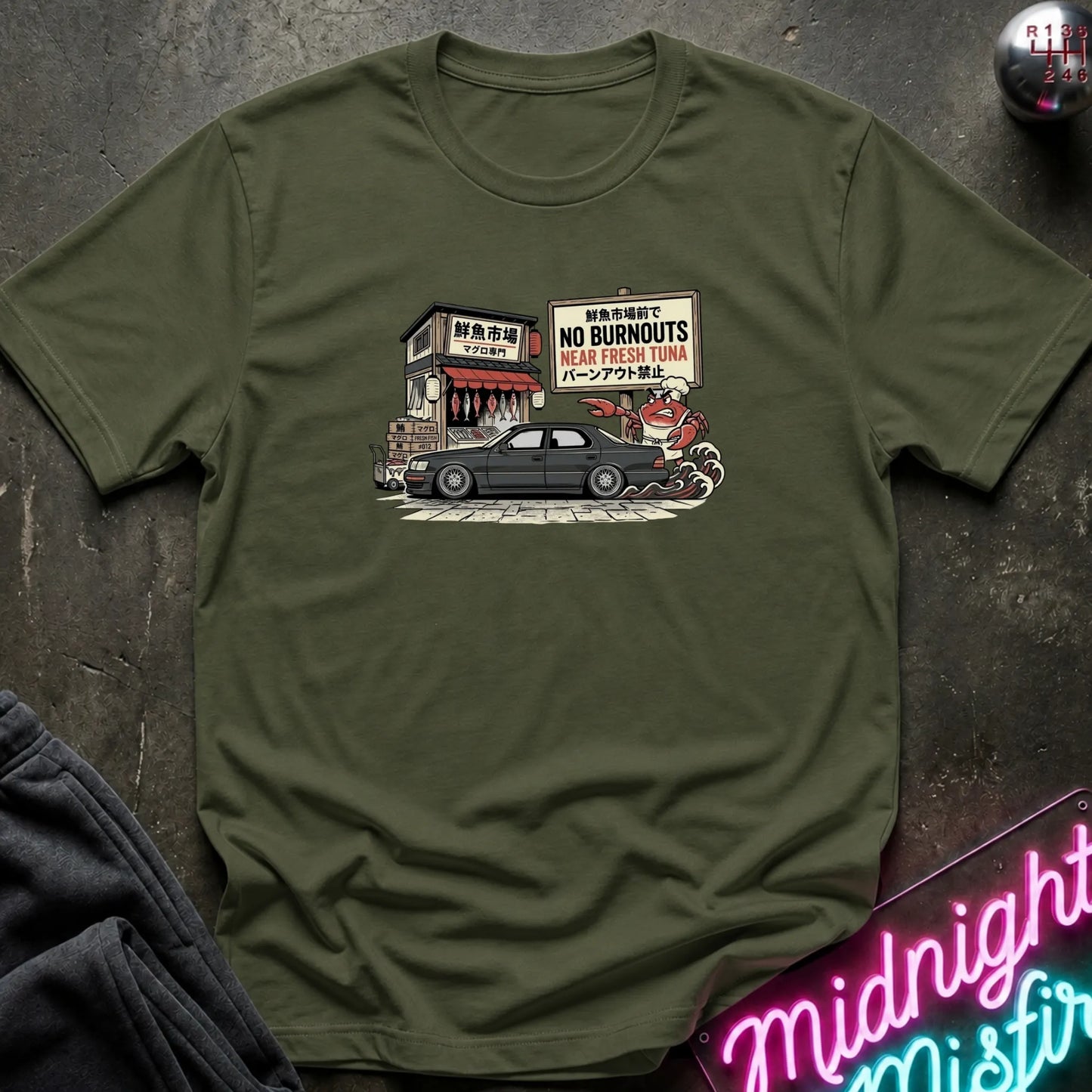 An olive green short-sleeve t-shirt with a graphic print of a black car parked in front of a Japanese-style restaurant with a 'No Burnouts' sign, laid flat on a dark textured surface with a neon sign and dark pants partially visible.