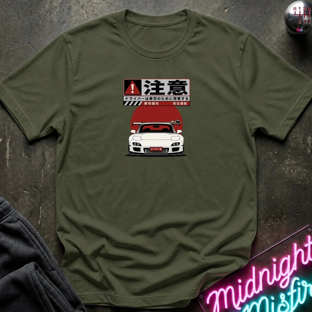 An olive green crewneck t-shirt featuring a graphic print of a white sports car and Japanese text, styled flat on a dark textured background with a neon sign and dark pants partially visible.