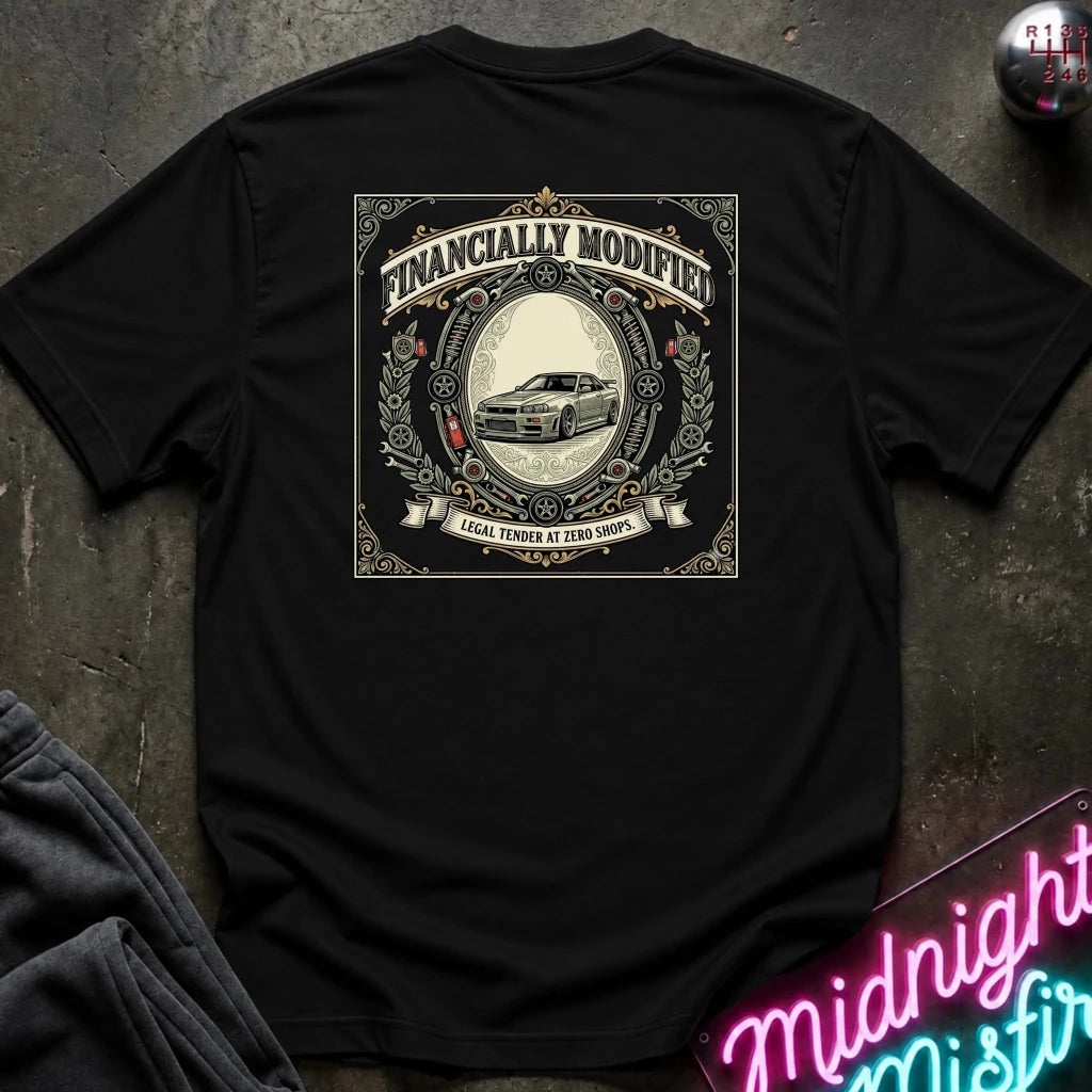 A black short-sleeve t-shirt laid flat on a dark textured surface, showing a large vintage-style graphic on the back with the words 'FINANCALLY MODIFIED' and a car illustration. A neon sign and folded dark fabric are partially visible around the shirt.
