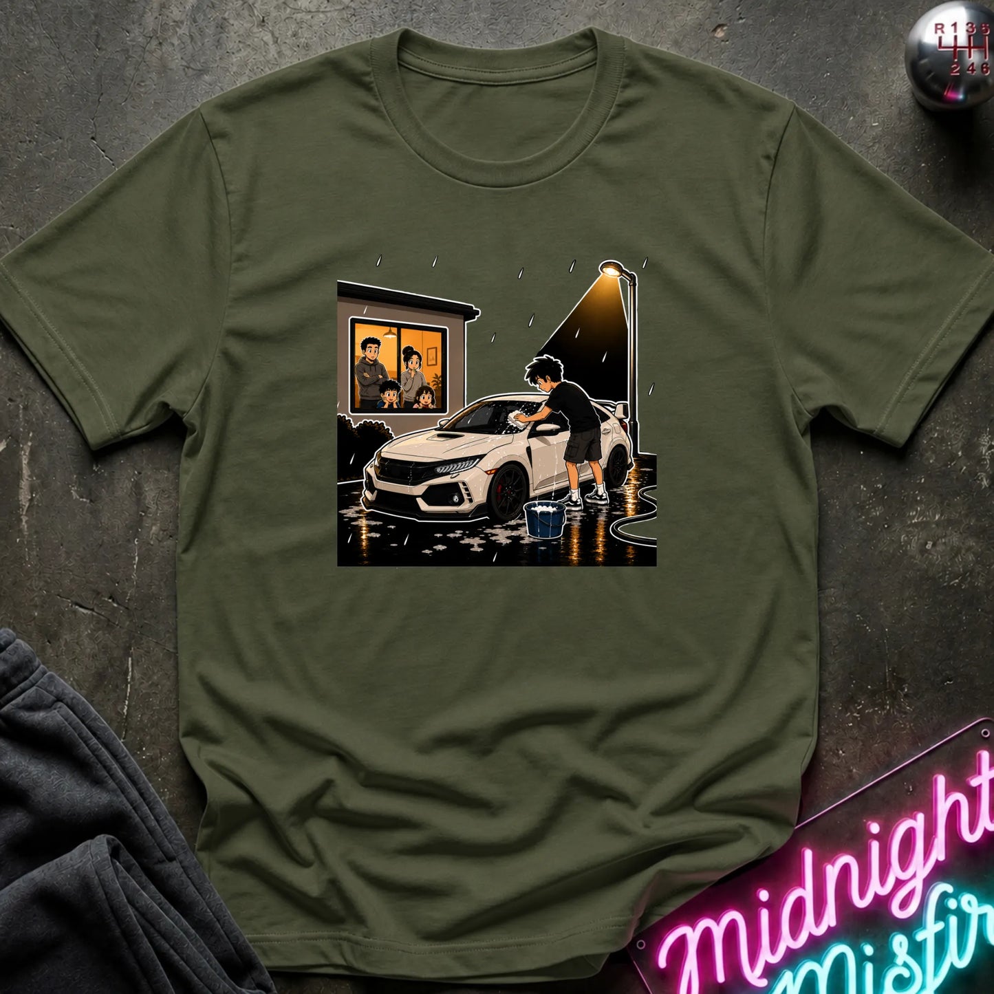An olive green short-sleeve t-shirt with a graphic print of a person washing a white car at night, displayed flat on a dark textured surface with a neon sign and dark sweatpants in the background.