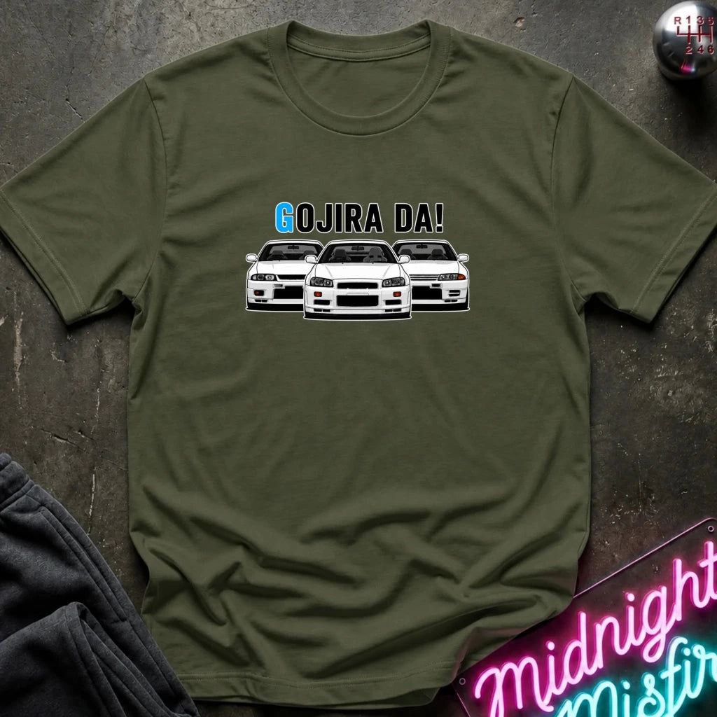 An olive green crewneck t-shirt featuring a graphic of three white Nissan Sentra cars with the text 'GOJIRA DA!' printed above them. The shirt is displayed flat on a dark textured surface with a neon sign and dark sweatpants partially visible.