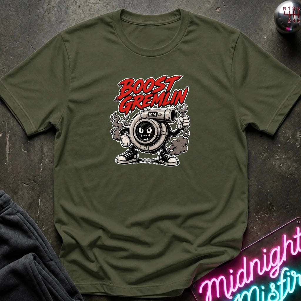 An olive green crewneck t-shirt featuring a cartoon graphic of a gas pump character with the text 'BOOST GREMLIN' in red and white, styled flat on a dark textured background with a neon sign and sweatpants partially visible.