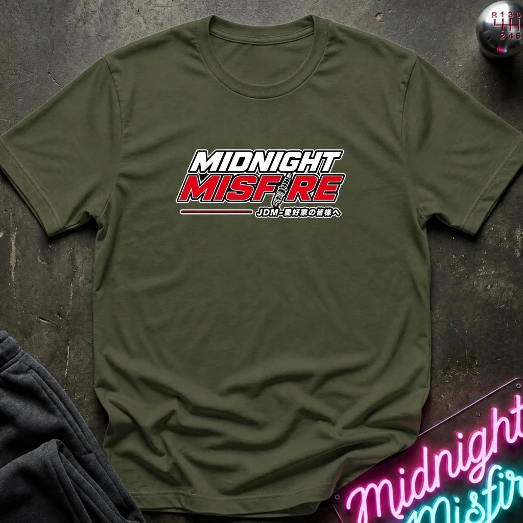 An olive green short-sleeve t-shirt laid flat on a dark textured surface, featuring a 'MIDNIGHT MISFIRE' graphic on the chest with stylized white and red lettering. A neon sign and folded dark clothing are partially visible in the background as props.