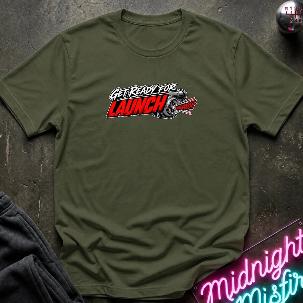 An olive green crewneck t-shirt with a 'GET READY FOR LAUNCH' graphic featuring a red and white rocket and a black snake, laid flat on a dark textured surface with a neon sign and dark pants in the background.