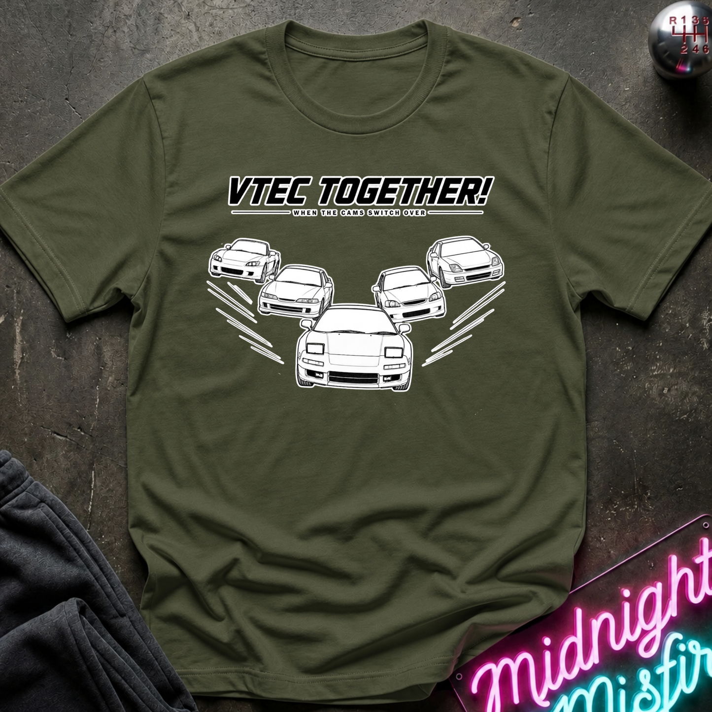 An olive green crewneck t-shirt featuring a white graphic of several sports cars with the text 'VTEC TOGETHER!, printed on the chest. The shirt is displayed flat on a dark textured surface with a neon sign and dark sweatpants partially visible.