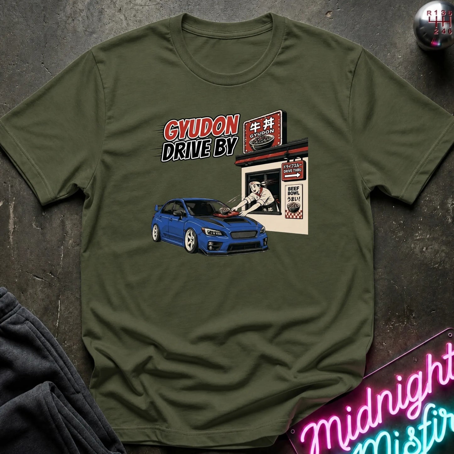 An olive green crewneck t-shirt laid flat on a dark textured surface, featuring a graphic print of a blue Subaru car and a Japanese restaurant with the text 'GYUDON DRIVE BY'.