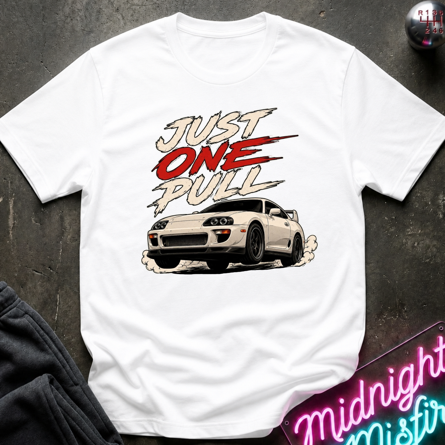 A white short-sleeve graphic t-shirt laid flat on a dark textured surface. The shirt features a graphic of a white sports car with the text 'JUST ONE PULL' in stylized red, black, and white lettering.