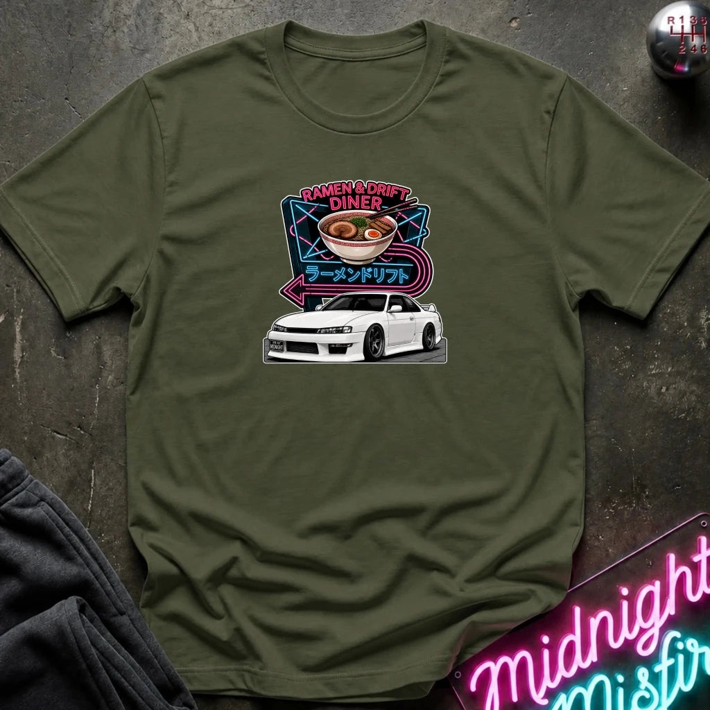 An olive green short-sleeve t-shirt with a graphic print of a white sports car and ramen bowl with neon-style text, laid flat on a dark textured surface with a neon sign and dark pants partially visible.