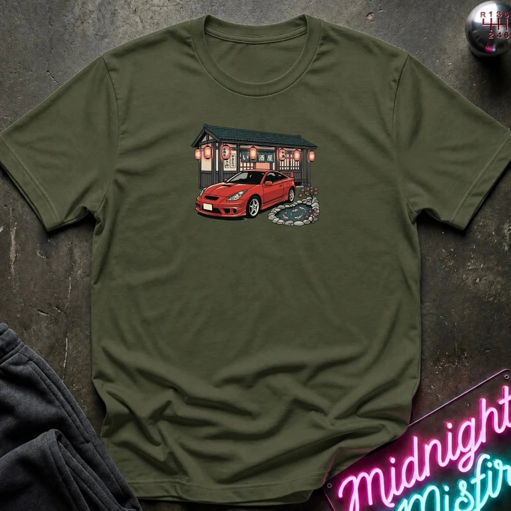 An olive green short-sleeve t-shirt laid flat on a dark textured surface, featuring a graphic print of a red sports car parked in front of a Japanese-style building with lanterns. A neon sign and folded dark fabric are partially visible as props.