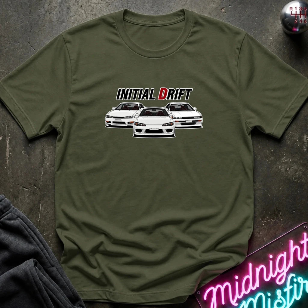An olive green short-sleeve t-shirt laid flat on a dark textured surface, featuring a graphic print of three white cars with the text 'INITIAL DRIFT' above them. A neon sign and folded dark fabric are partially visible around the shirt.