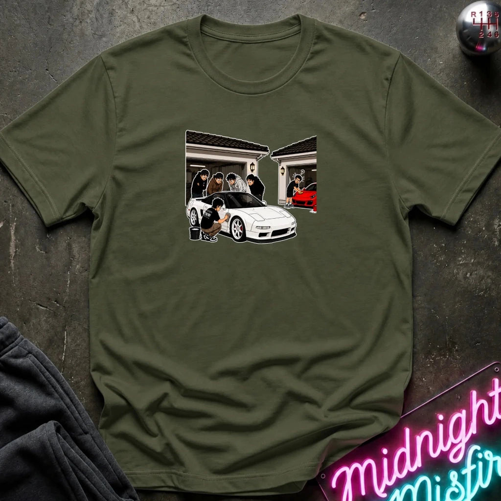 An olive green short-sleeve t-shirt with a graphic print of a white sports car in a garage, laid flat on a dark textured surface with a neon sign and dark pants partially visible.