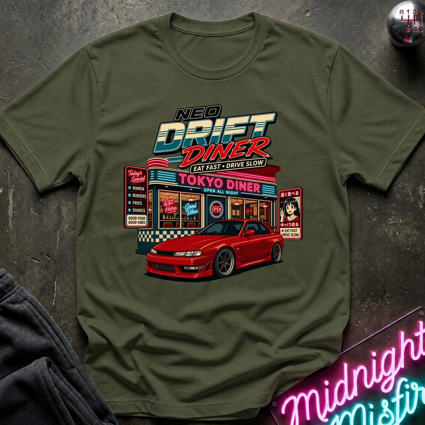 An olive green crewneck t-shirt featuring a colorful graphic print of a 'Neo Drift Diner' with a red sports car in front, laid flat on a dark textured surface with a neon sign and dark fabric in the corners.