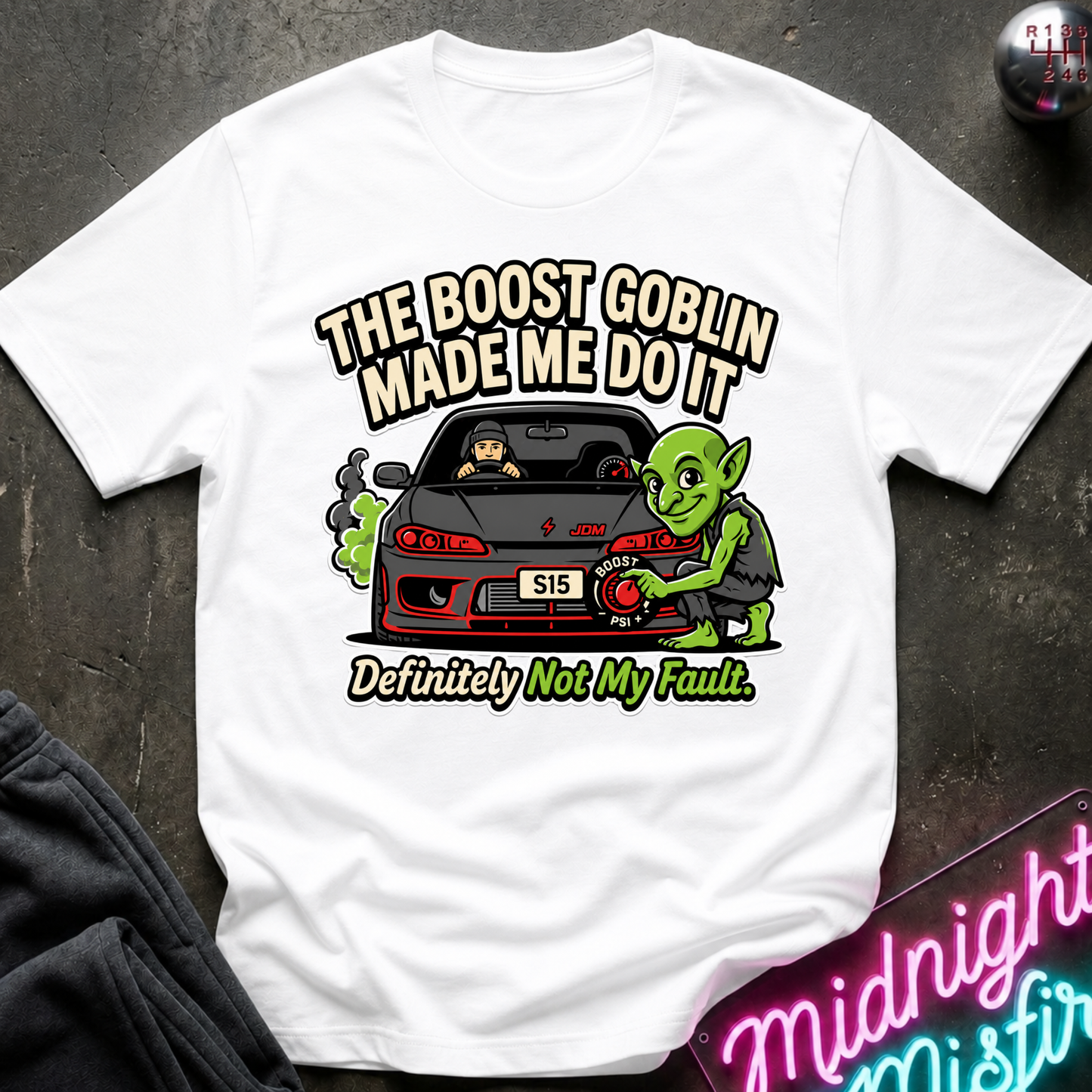 A white short-sleeve graphic T-shirt laid flat on a dark textured surface. The shirt features a cartoon illustration of a green goblin working on a black car with the text 'THE BOOST GOBLIN MADE ME DO IT' above and 'Definitely Not My Fault.