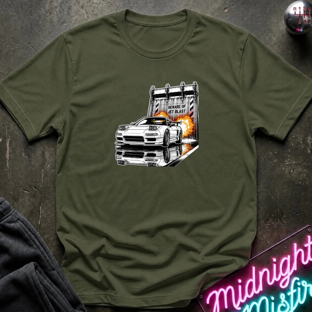 An olive green  short-sleeve t-shirt laid flat on a dark textured surface, featuring a graphic print of a white sports car in front of a blast off area.