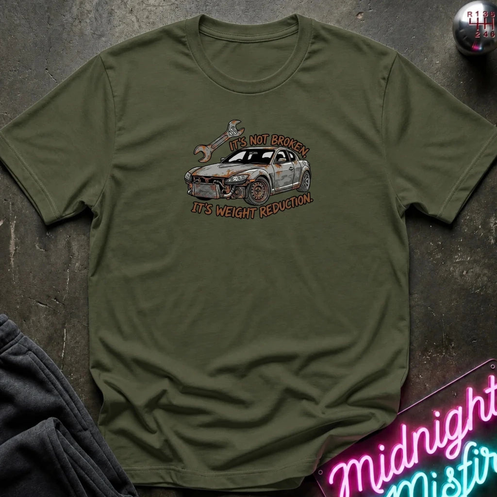 An olive green crewneck t-shirt with a graphic of a white and gray sports car and the text 'IT'S NOT BROKEN IT'S WEIGHT REDUCTION' in a distressed orange font. The shirt is laid flat on a dark textured surface with a neon sign and dark pants partially visible.
