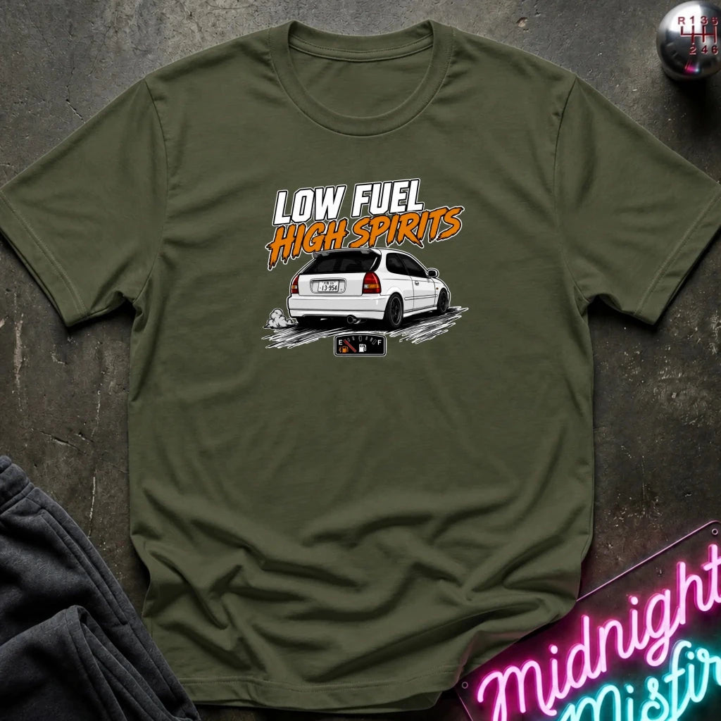 An olive green short-sleeve t-shirt laid flat on a dark textured surface. The shirt features a graphic print of a white car with the text 'LOW FUEL HIGH SPIRITS' in white and orange.