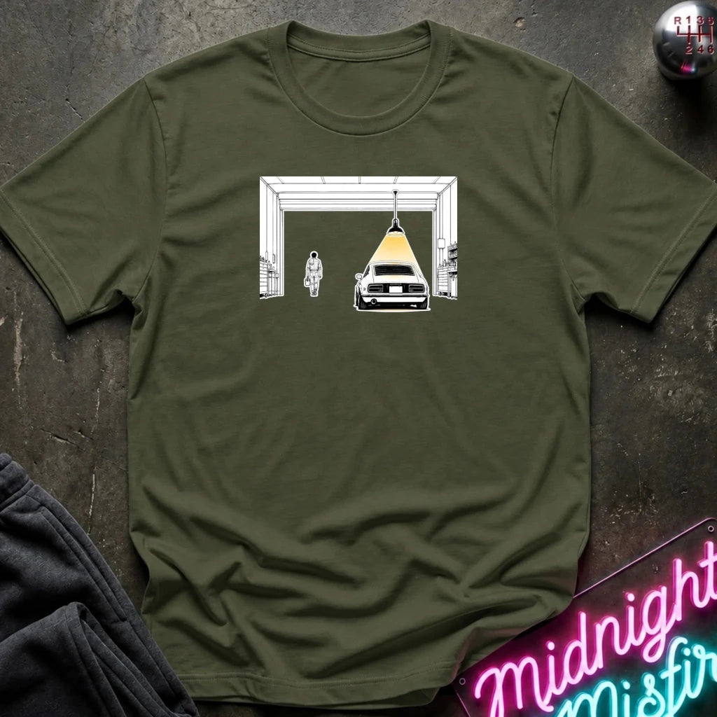 An olive green crewneck t-shirt laid flat on a dark textured surface, featuring a graphic print of a white car under a yellow spotlight inside a white frame. 
