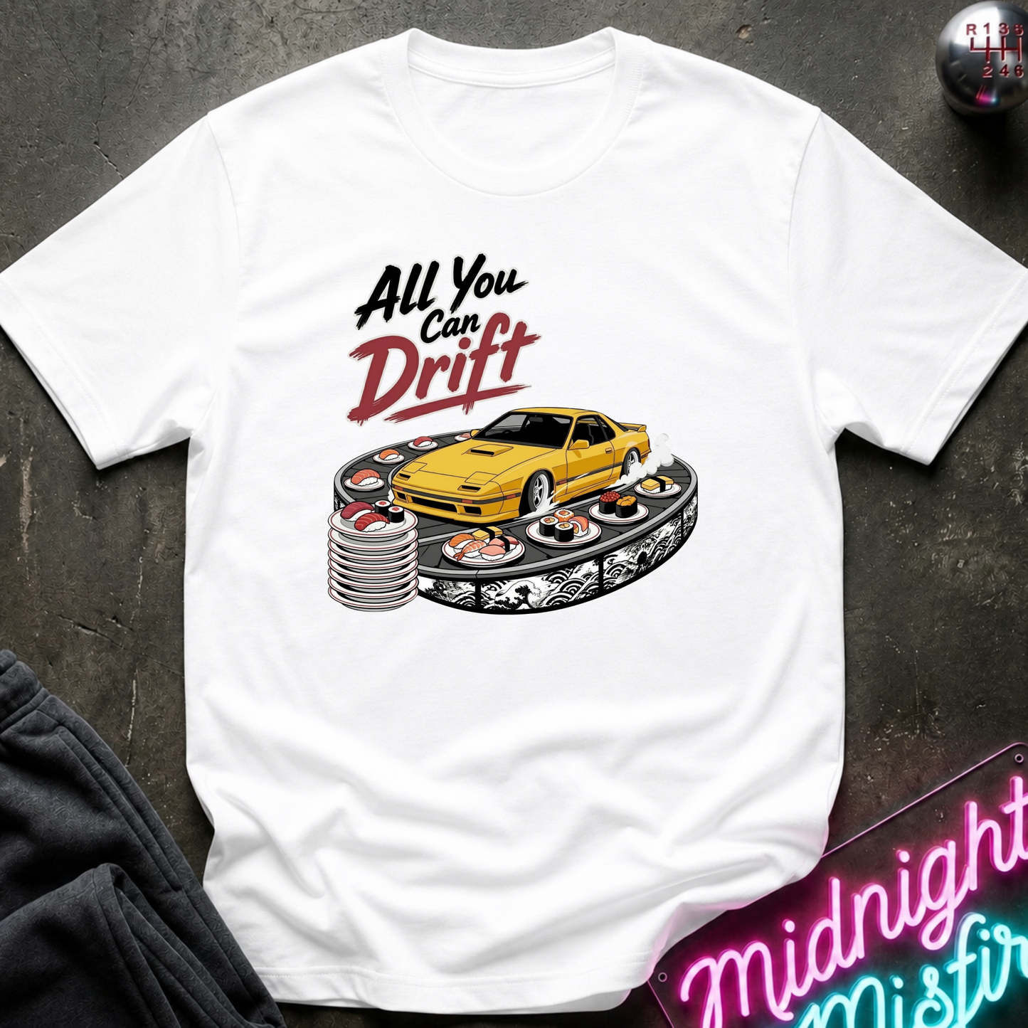 A white short-sleeve graphic t-shirt laid flat on a dark textured surface. The graphic features the text 'All You Can Drift' in black and red lettering above a yellow sports car drifting on a track with sushi plates.