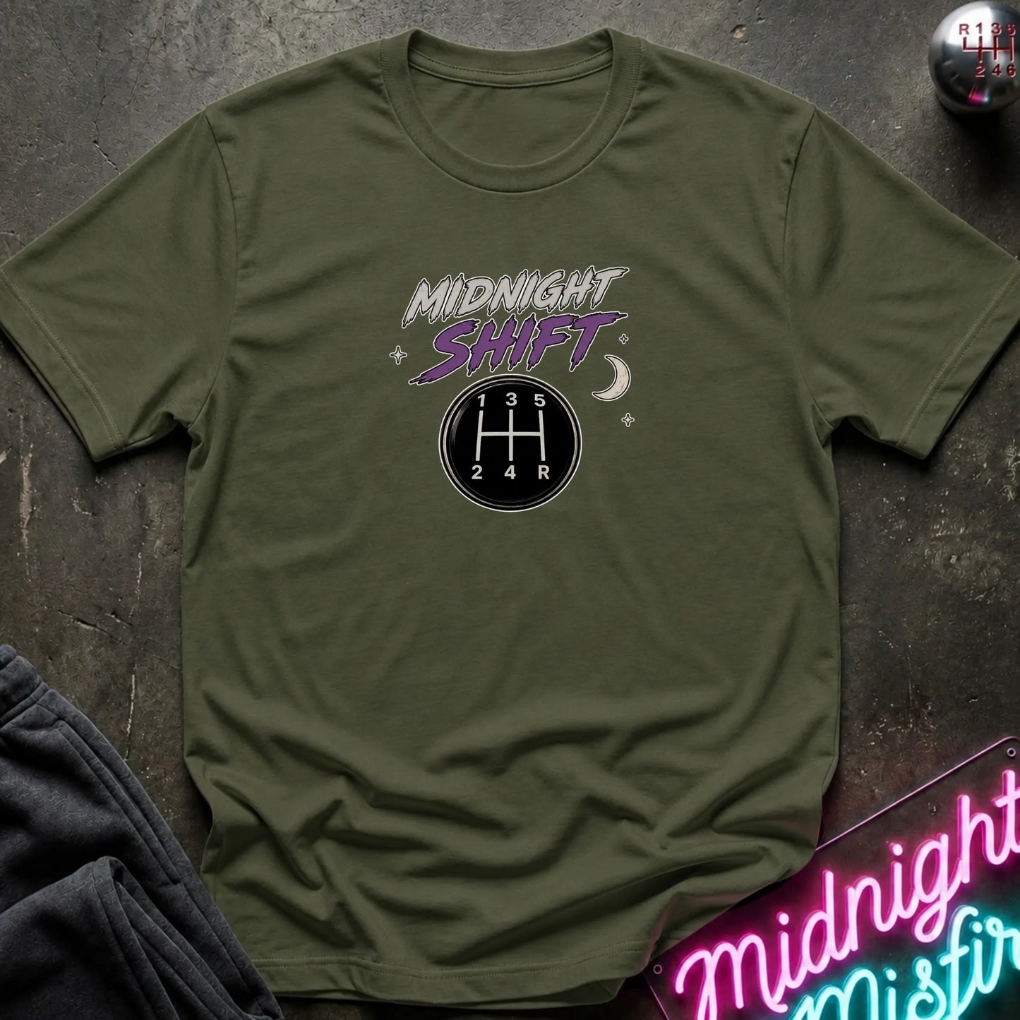 An olive green crewneck t-shirt laid flat on a dark textured surface, featuring a 'Midnight Shift' graphic with a 5-speed manual gearshift pattern and stars. A neon sign and dark clothing are partially visible as props.
