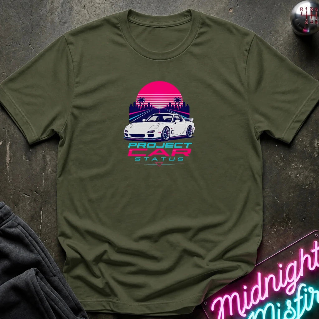 An olive green short-sleeve t-shirt laid flat on a dark textured surface, featuring a retro-style graphic of a white sports car in front of a pink and purple sunset with palm trees and the text 'PROJECT CAR STATUS'.