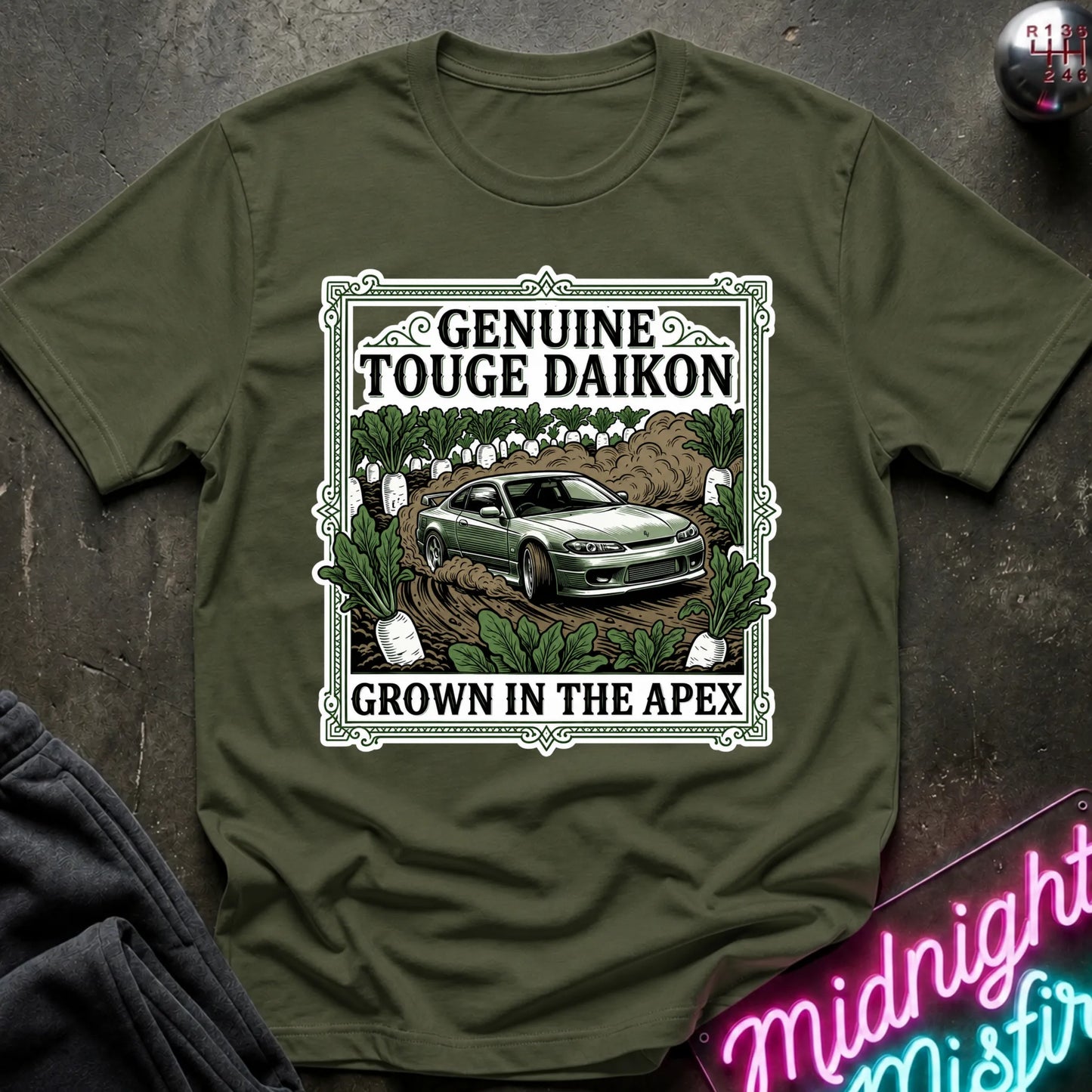 An olive green short-sleeve graphic T-shirt laid flat on a dark textured surface. The shirt features a large rectangular front print with the words 'GENUINE TOUGHE DAIKON' and 'GROWN IN THE APEX'.