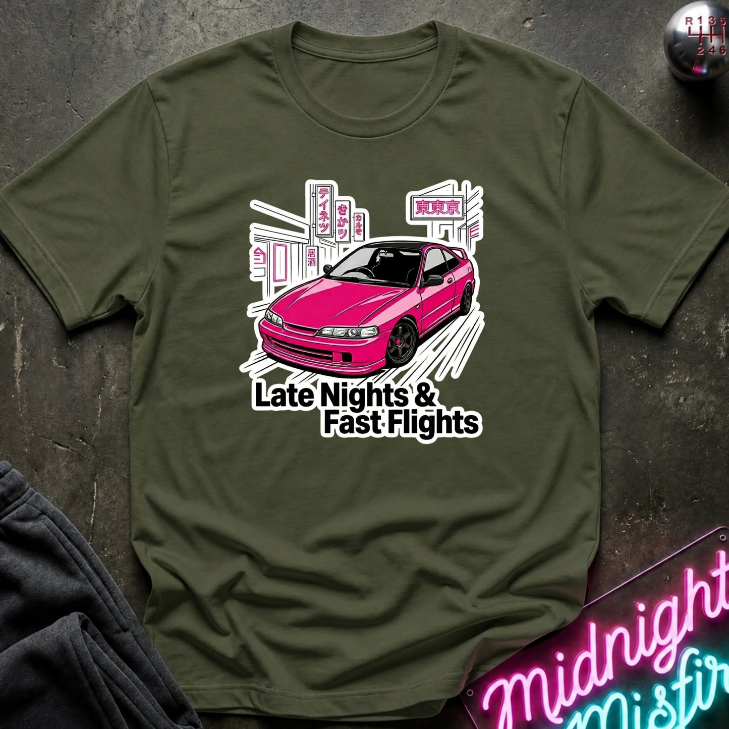An olive green crewneck t-shirt laid flat on a dark textured surface, featuring a graphic of a pink car with the text 'Late Nights & Fast Flights'.