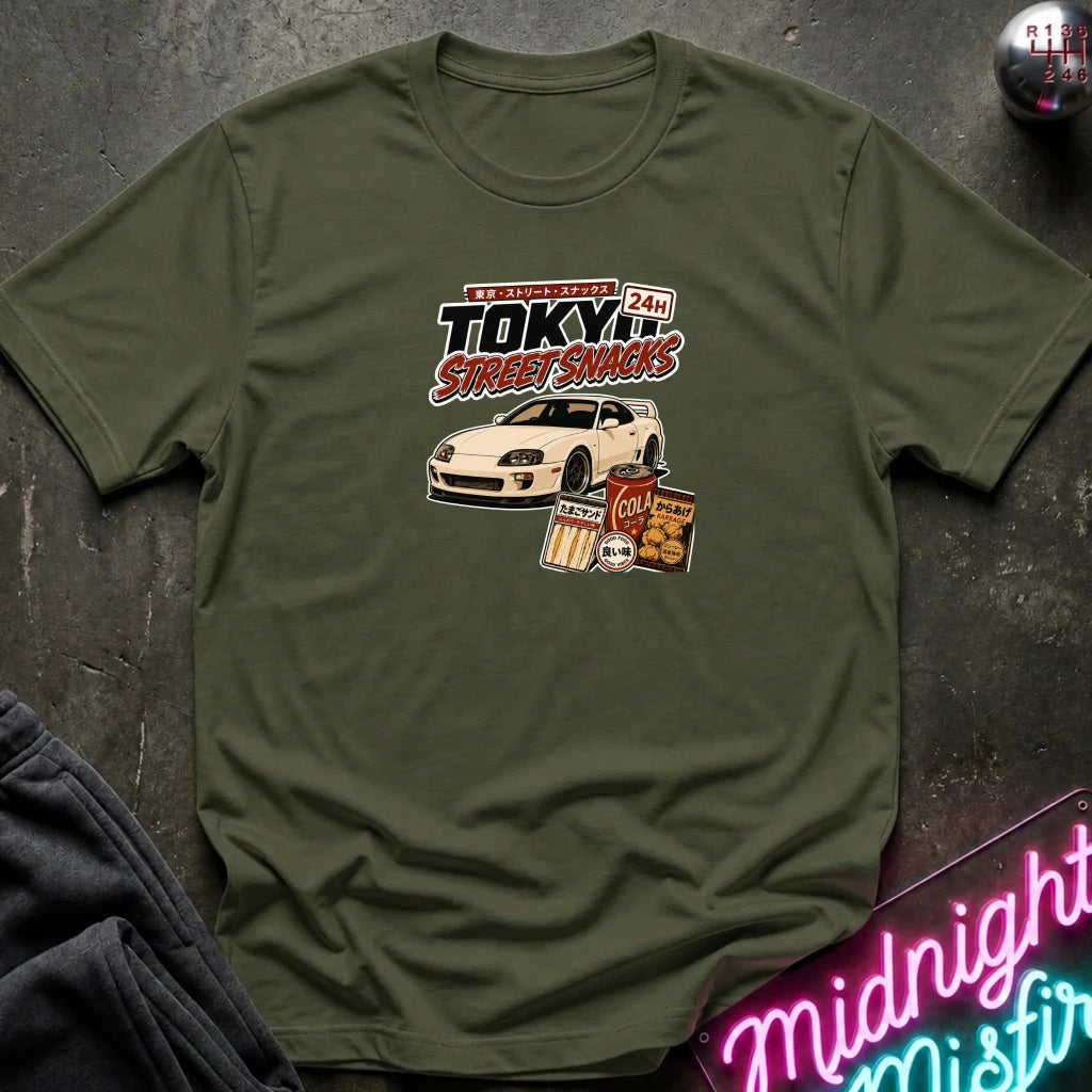 An olive green short-sleeve graphic T-shirt laid flat on a dark textured surface. The shirt features a central graphic with the text 'Tokyo Street Snacks' above a white sports car and snack illustrations.