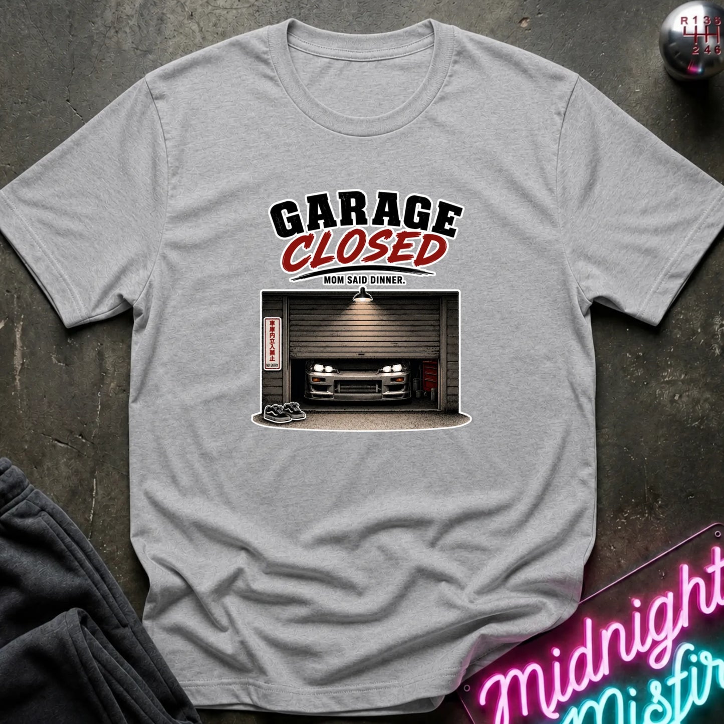 A light gray heather crewneck t-shirt laid flat on a dark textured surface. The shirt features a graphic print with the text 'GARAGE CLOSED' in black and red, above an illustration of a car parked inside a closed garage.