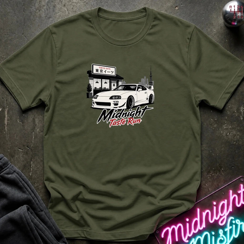 An olive green short-sleeve t-shirt laid flat on a dark textured surface, featuring a graphic print of a white sports car in front of a building with the text 'Midnight Taste Run'.