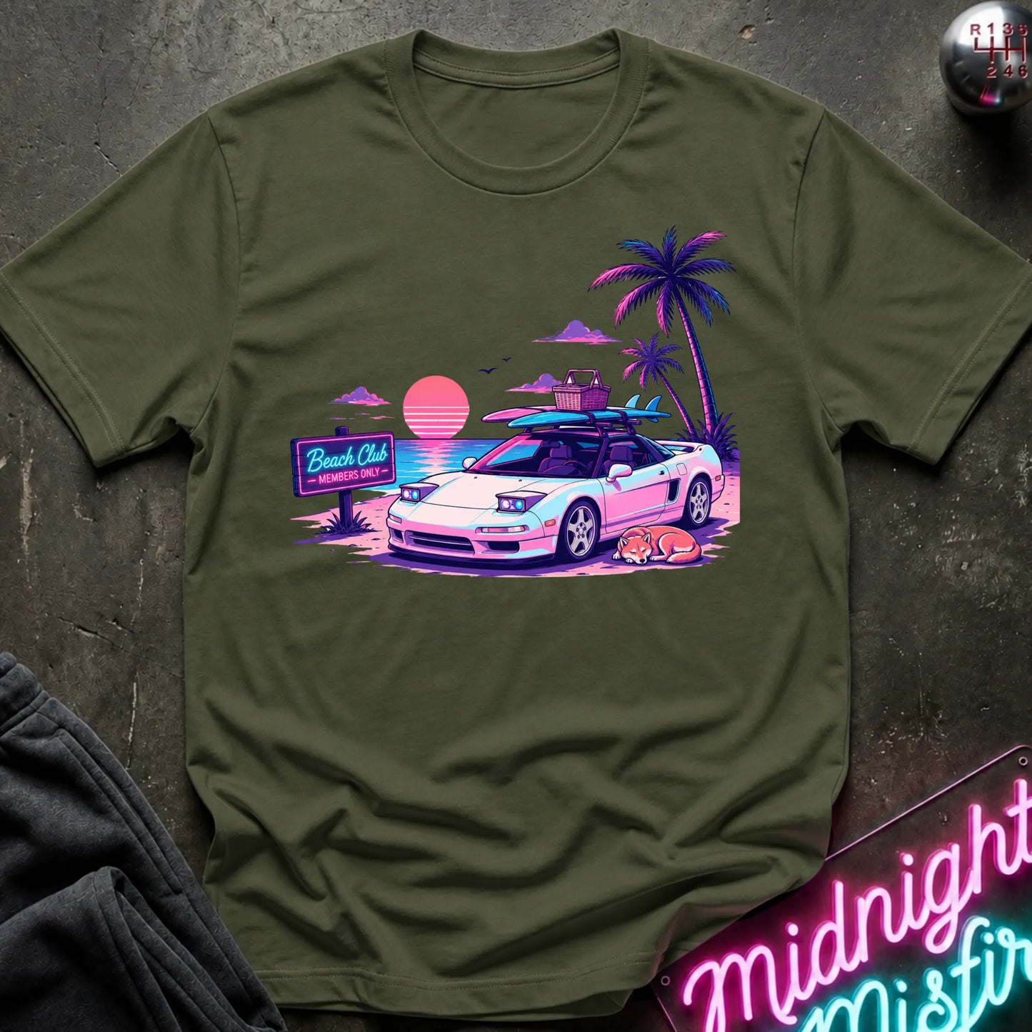 An olive green short-sleeve graphic t-shirt laid flat on a dark textured surface. The graphic features a retro-style illustration of a sports car with surfboards on its roof, next to palm trees, a sunset, and a 'Beach Club' sign.