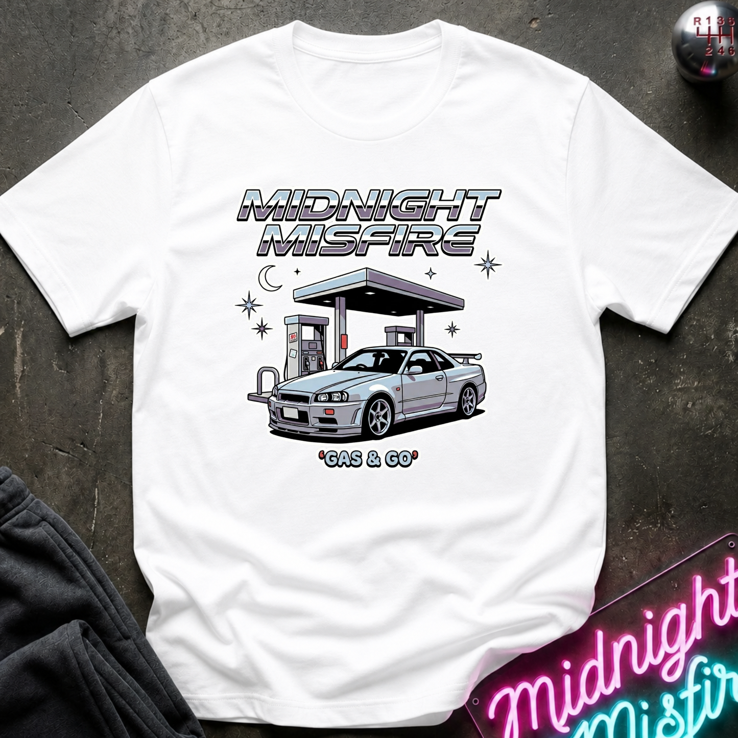 A white short-sleeve graphic T-shirt laid flat on a dark textured surface. The shirt features a printed design of a silver sports car at a gas station with the text 'MIDNIGHT MISFIRE' and '*GAS & GO'.