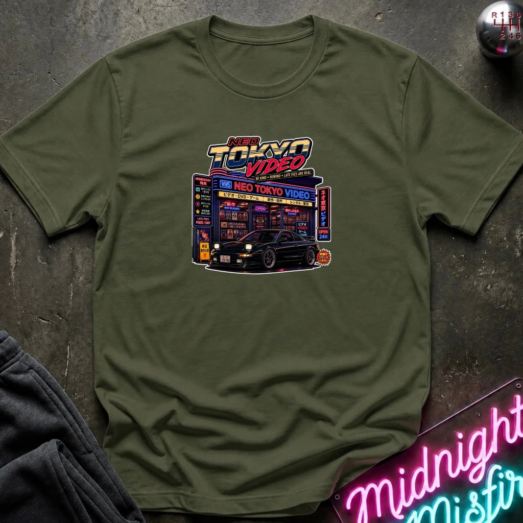 An olive green short-sleeve t-shirt with a colorful graphic print of a 'Neo Tokyo Video' arcade and a black sports car, styled flat on a dark textured surface with a neon sign and dark pants in the background.