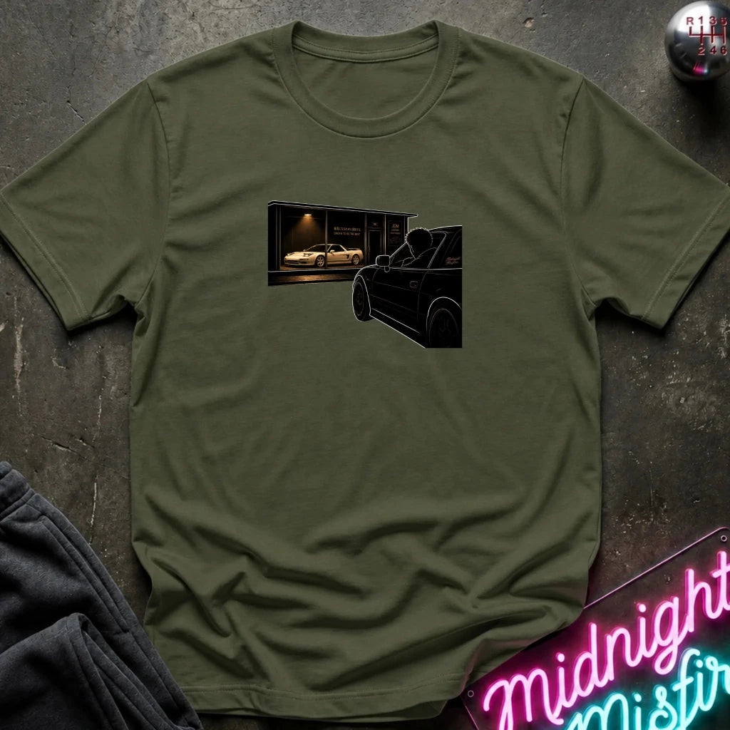 An olive green crewneck t-shirt laid flat on a dark textured surface, featuring a graphic print of a black car silhouette in front of a garage with a white car inside.