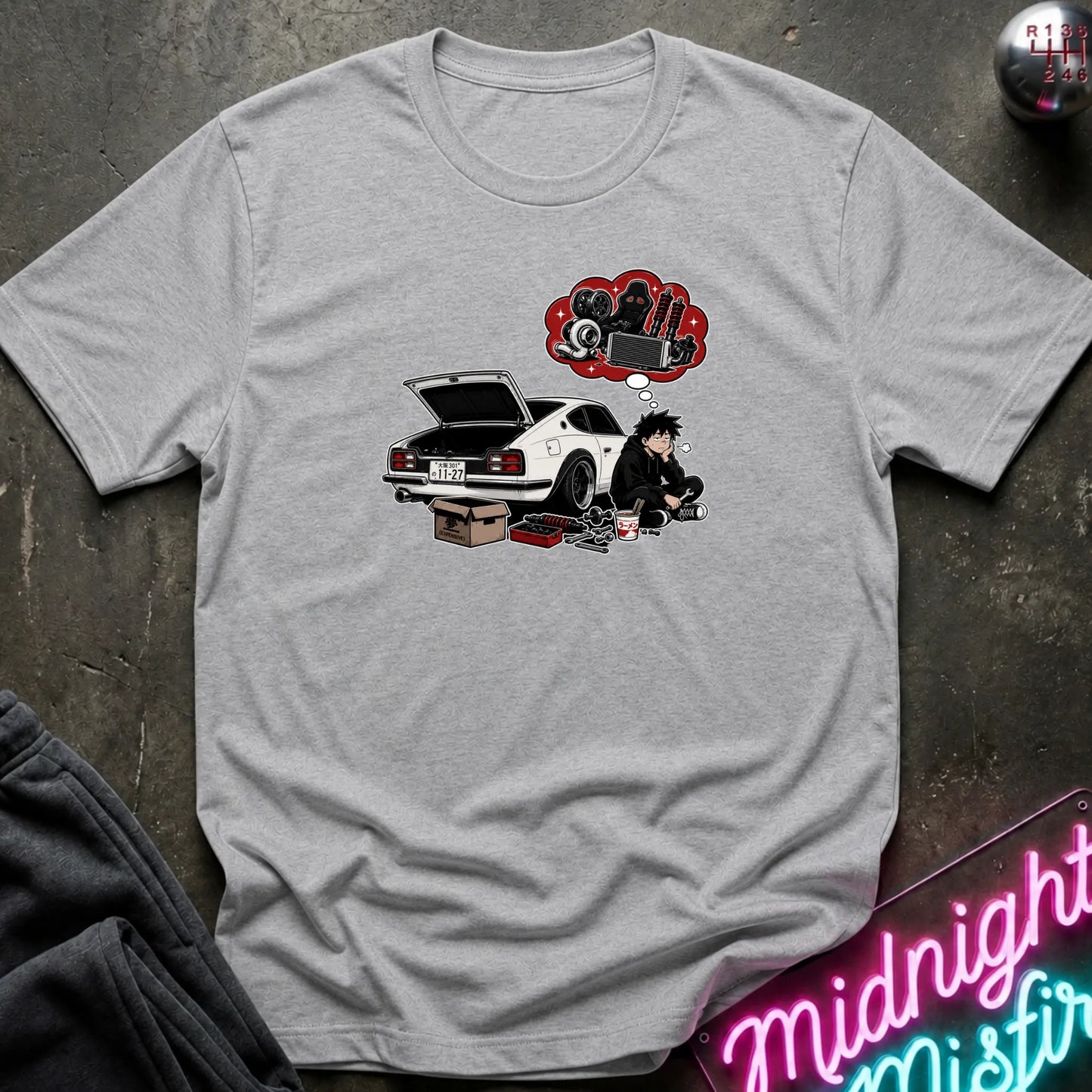 A light gray heather  short-sleeve graphic t-shirt laid flat on a dark textured surface. The graphic features a white sports car with its trunk open, a cartoon character sitting beside it, and a red thought bubble filled with car parts.