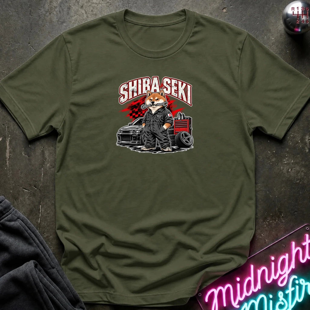 An olive green crewneck t-shirt featuring a graphic of a fox in a suit standing next to a car with the text 'SHIBA SEKI' above it, laid flat on a dark textured surface with a neon sign and dark pants partially visible.