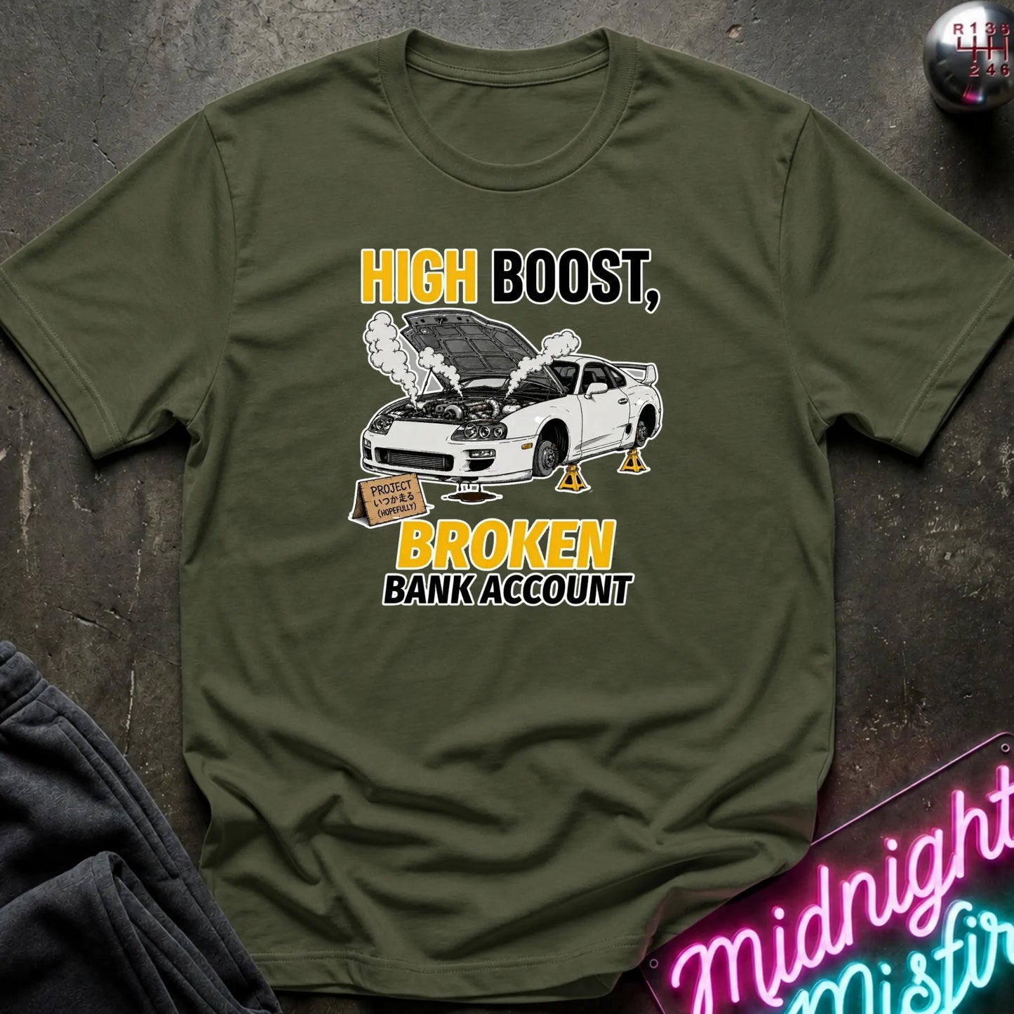 An olive green graphic t-shirt laid flat on a dark textured surface. The graphic features a white sports car with its hood open, smoke, and a sign, with the text 'HIGH BOOST, BROKEN BANK ACCOUNT' in yellow and black.