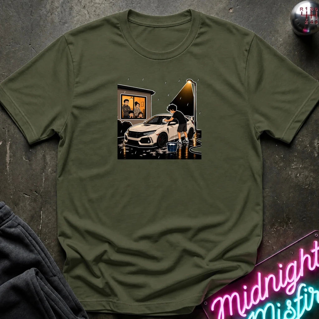 An olive green crewneck t-shirt laid flat on a dark textured surface, featuring a central graphic print of a white sports car in a rainy, neon-lit scene. 