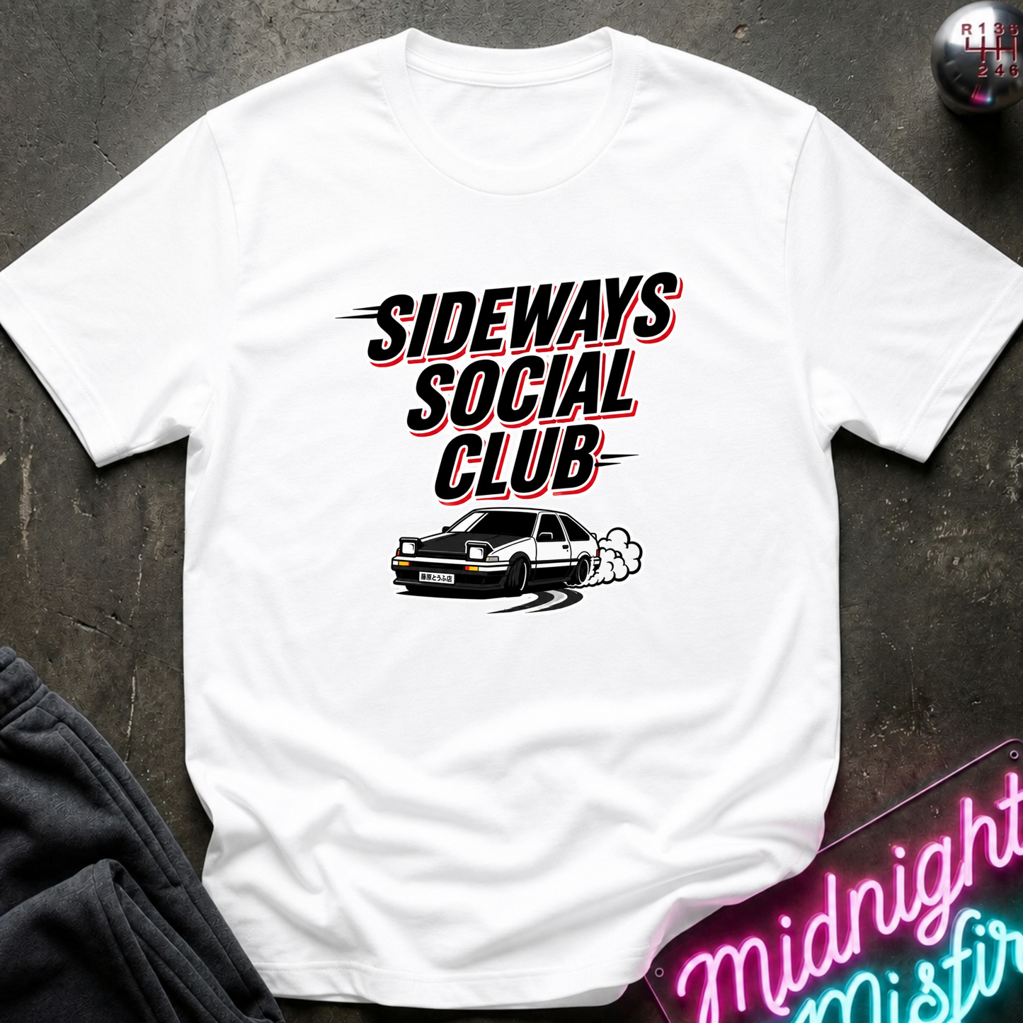 A white short-sleeve graphic T-shirt laid flat on a dark textured surface. The shirt features the text 'SIDEWAYS SOCIAL CLUB' in black with red and pink outline accents, above a black-and-white car graphic.