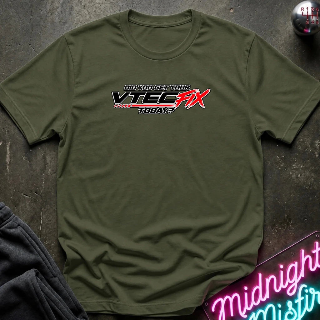 An olive green crewneck t-shirt laid flat on a dark textured surface, featuring a graphic print that reads 'dID YOU GET YOUR VITEC FIX TODAY?' in black and red. A pair of dark sweatpants is partially visible in the bottom-left corner, and a neon sign is in the bottom-right corner.