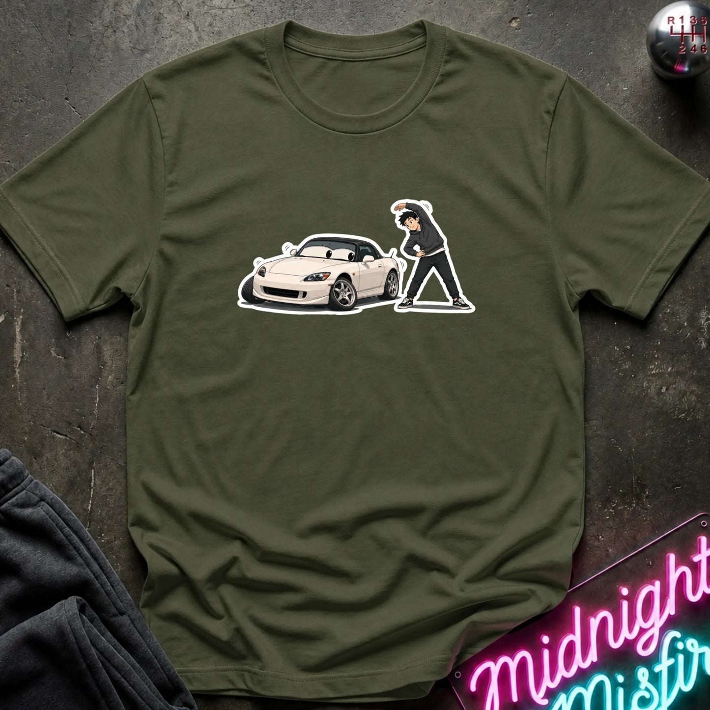 An olive green short-sleeve t-shirt with a graphic print of a white sports car and a s figure, laid flat on a dark textured surface with a neon sign and dark pants partially visible.