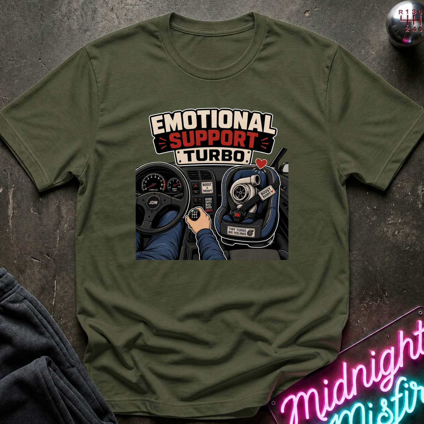 An olive green graphic t-shirt laid flat on a dark textured surface. The shirt features a printed graphic of a car dashboard and steering wheel with the text 'EMOTIONAL SUPPORT TURBO' in white and red.