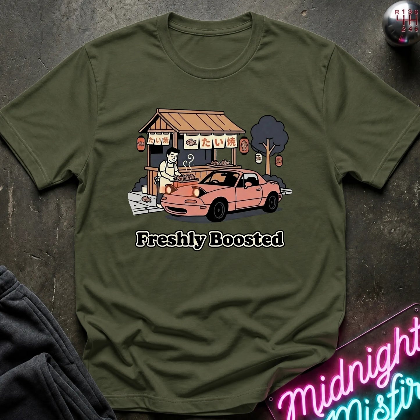 An olive green graphic t-shirt laid flat on a dark textured surface. The shirt features a cartoon illustration of a street food stall with a pink car and the text 'Freshly Boosted'.