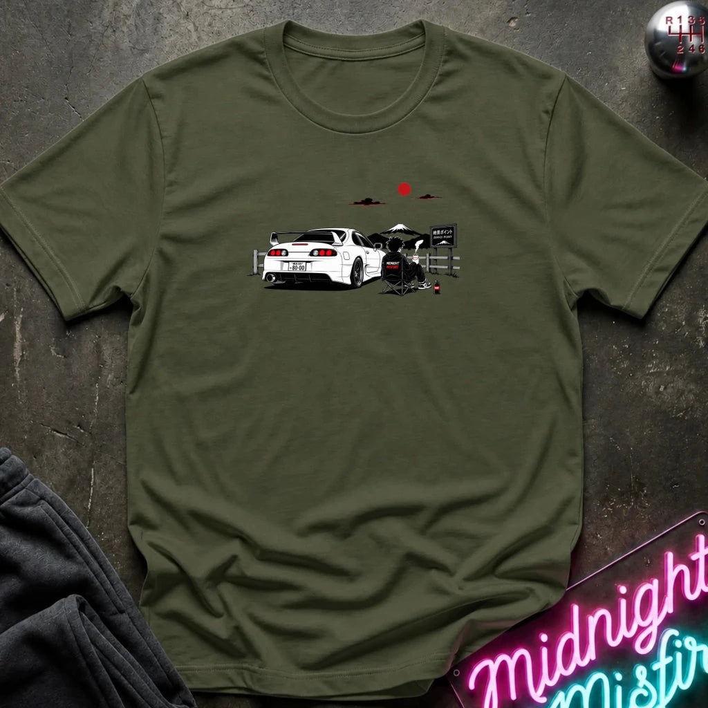 An olive green short-sleeve graphic T-shirt laid flat on a dark textured surface. The shirt features a printed illustration of a white sports car with a red sun and mountain graphic above it.