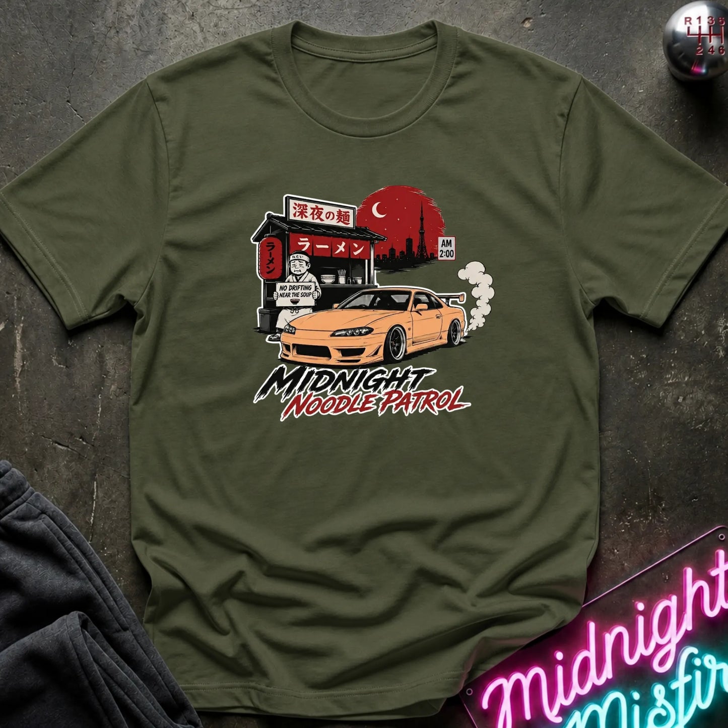 An olive green crewneck t-shirt laid flat on a dark textured surface, featuring a graphic print of a yellow sports car in front of a ramen shop at night with the text 'MIDNIGHT NOODLE PATROL'.