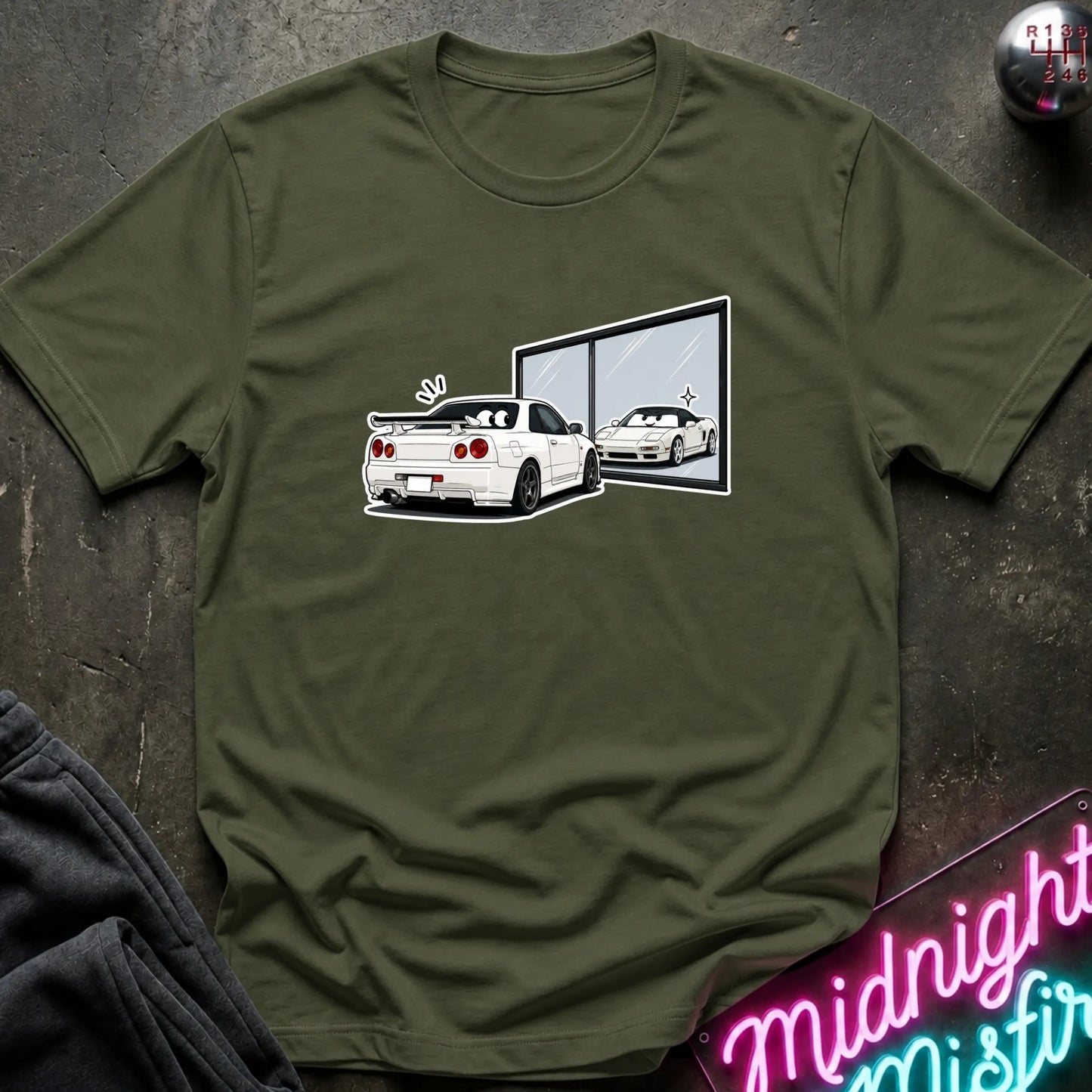 An olive green short-sleeve t-shirt laid flat on a dark textured surface, featuring a graphic print of a white sports car and its reflection in a window. A neon sign and dark clothing are partially visible around the edges.