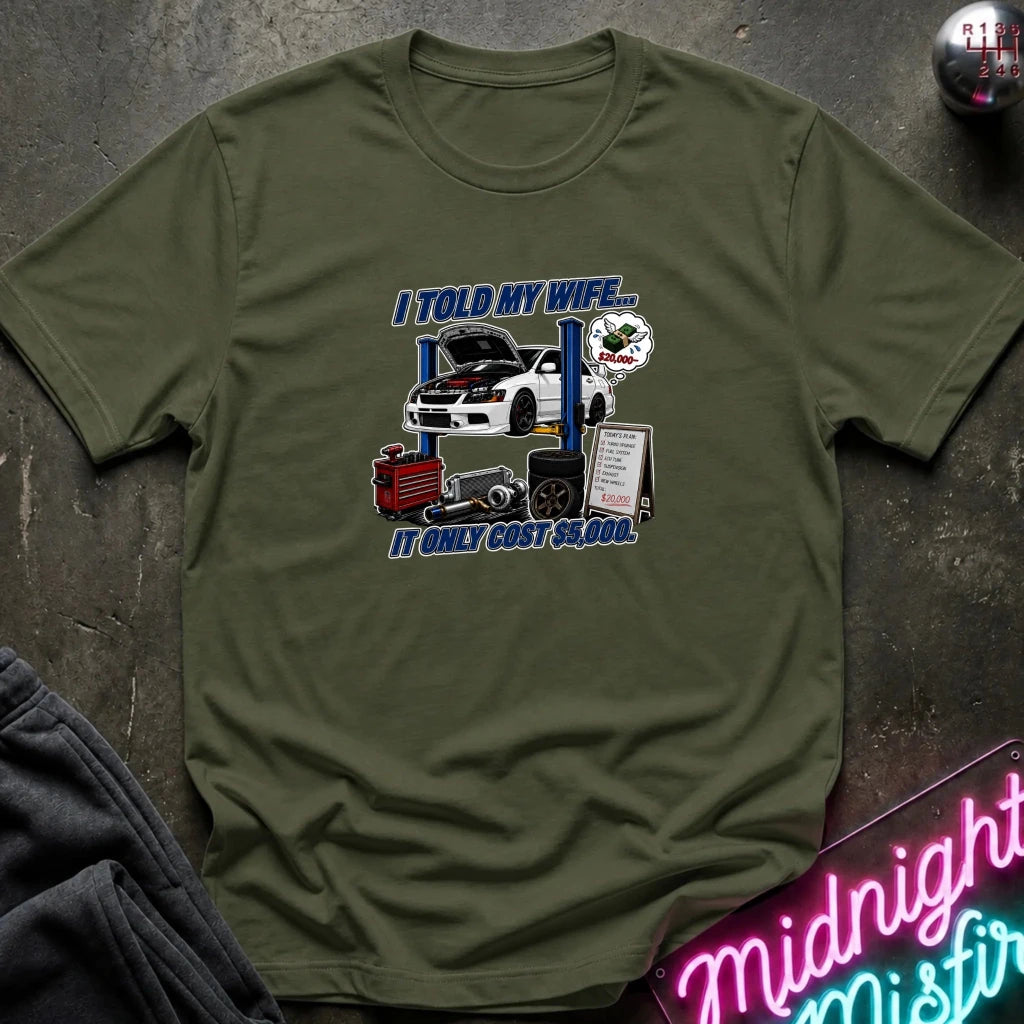 An olive green short-sleeve t-shirt with a humorous car graphic printed on the chest, laid flat on a dark textured surface with a neon sign and dark pants partially visible.