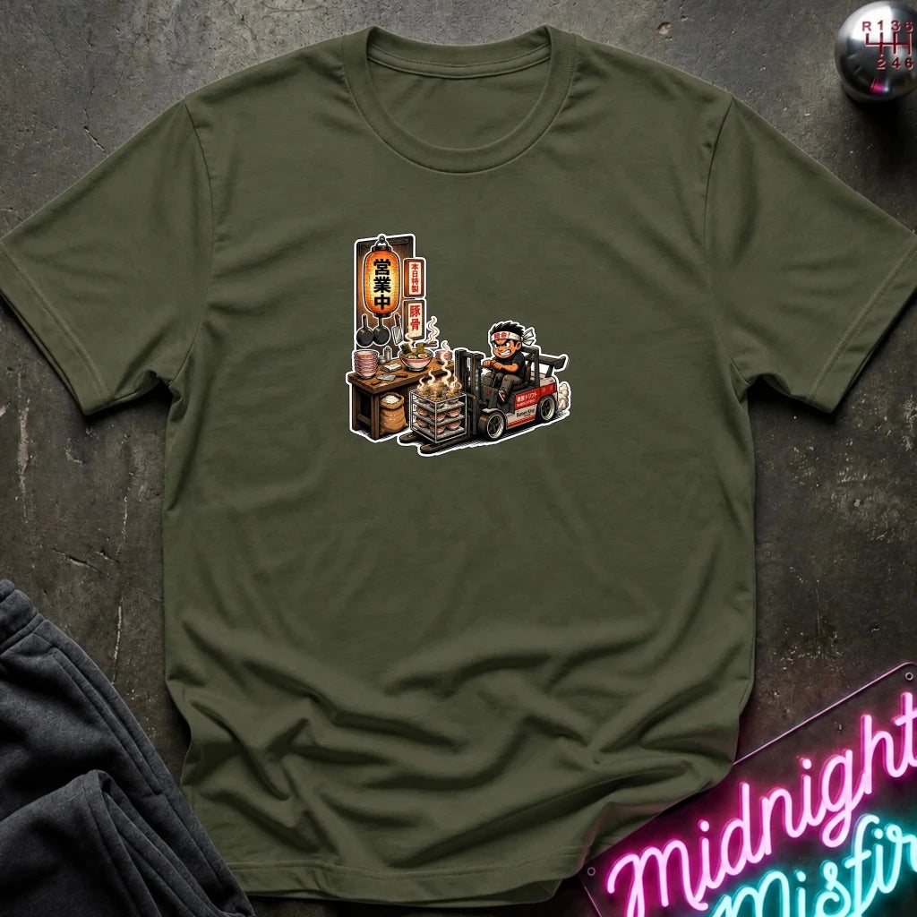 An olive green short-sleeve t-shirt with a cartoon graphic of a street food vendor on a forklift, laid flat on a dark textured surface with a neon sign and folded sweatpants nearby.