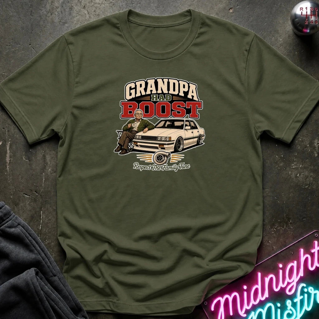 olive green short-sleeve graphic t-shirt laid flat on a dark textured surface. The graphic features the text 'GRANDPA RAW BOOST' in beige and red, above an illustration of a man sitting in a car with the phrase 'Respect The Family Tune' below.
