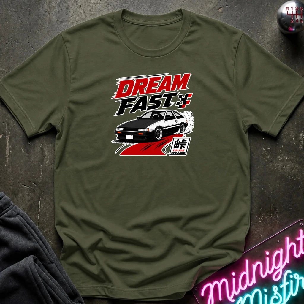 An olive green short-sleeve graphic T-shirt laid flat on a dark textured surface. The shirt features a red, black, and white 'DREAM FAST' graphic with a car illustration.