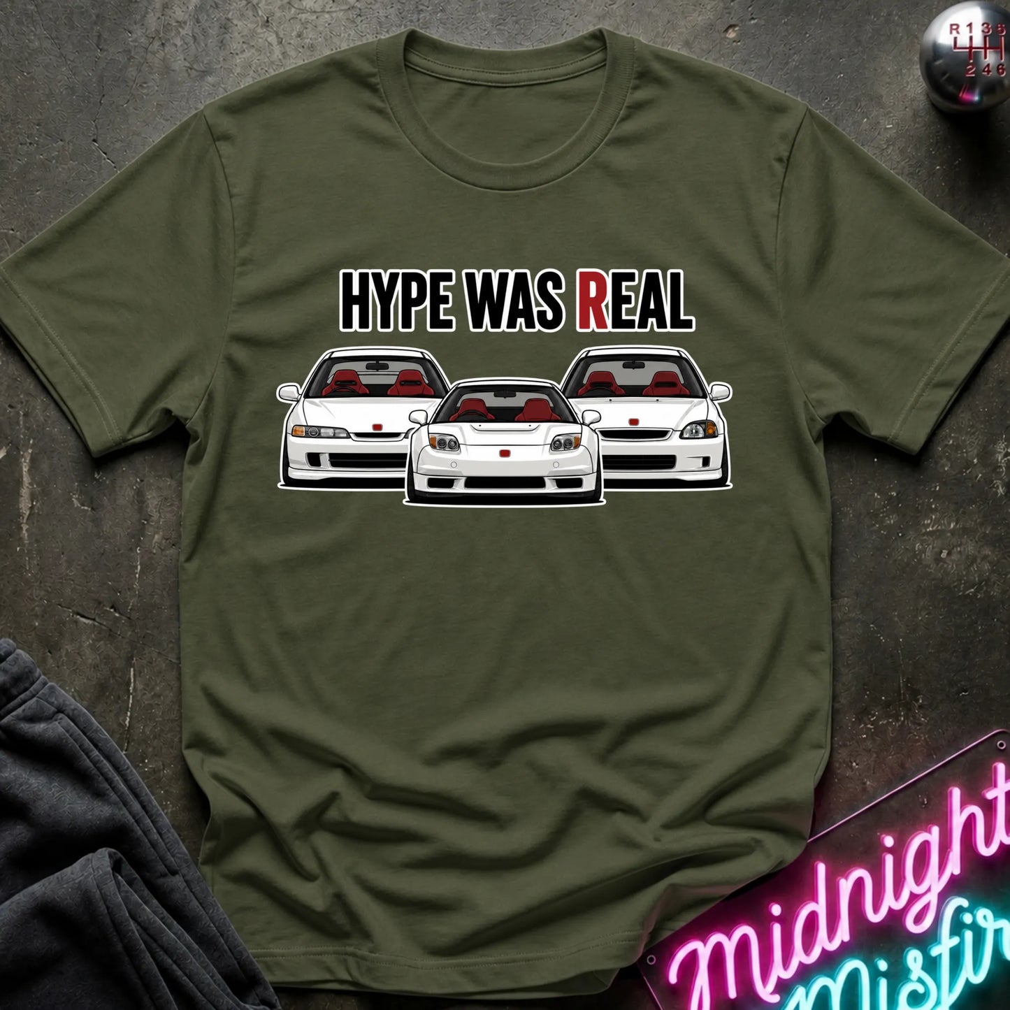 An olive green short-sleeve t-shirt with a graphic of three white sports cars and the text 'HYPE WAS REAL' in black and red, laid flat on a dark textured surface.
